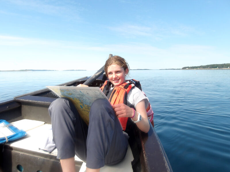 A young woman is sitting in a small boat on a calm body of water, possibly the ocean, under a clear blue sky. She is wearing a life jacket and holding a map, suggesting she might be navigating. The boat appears to be a simple, possibly homemade, rowboat. The background shows a distant shoreline with trees. The overall scene conveys a sense of adventure and peaceful exploration.